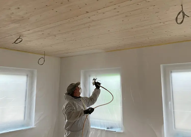 Foto eines Mitarbeiters der Malerei Norbert Bardtke beim Besprühen einer Holzdecke mit weißer Farbe. Die Wände des Raumes sind mit Plastikfolie behängt.