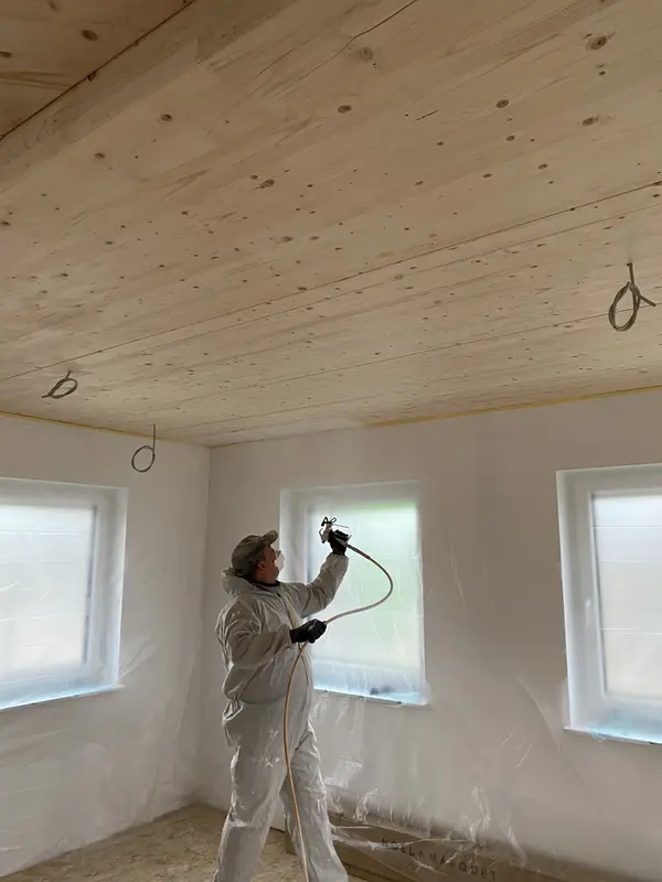 Foto eines Mitarbeiters der Malerei Norbert Bardtke beim Besprühen einer Holzdecke mit weißer Farbe. Die Wände des Raumes sind mit Plastikfolie behängt.