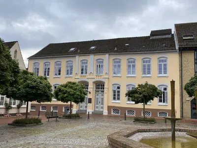 Foto eines von der Malerei Norbert Bardtke gelb gestrichenenen großen Hauses in einer Stadt. Vor dem Haus ist ein Platz mit SItzbänken, Pflanzen und einem Brunnen zu sehen.