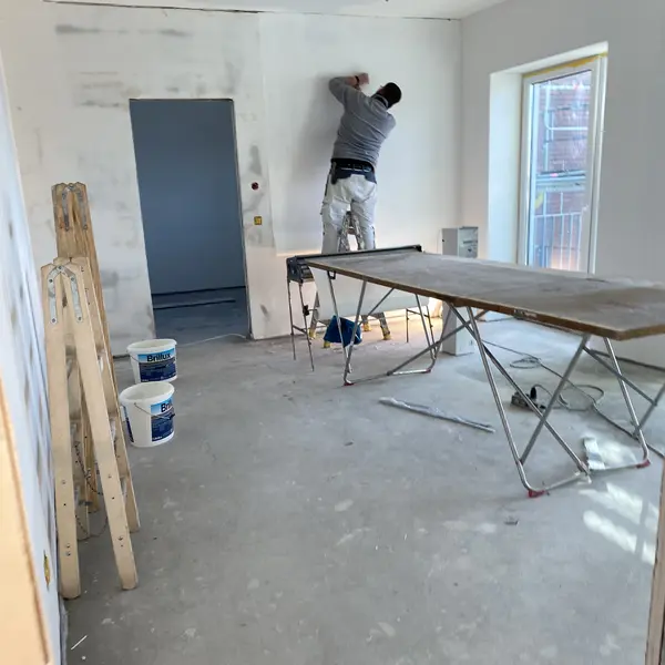 In einem Raum stehen ein Tapeziertisch, einige Holzleitern und Farbeimer auf dem Boden. Ein Mitarbeiter der Malerei Norbert Bardtke tapeziert eine Wand des Raumes mit einer hellen Tapete.