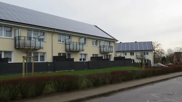Es sind zwei von der Malerei Norbert Bardtke gelb gestrichene Mehrfamilienhäuser an einem wolkenbedeckten Tag zu sehen. Die Häuser haben Solarpanele auf den Dächern und die Wände der Balkone bestehen aus Glas. 