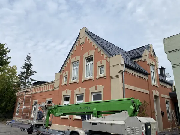 Ein rot gelbes Haus, vor welchem ein kleiner grüner Krahn mit der Aufschrift Malereibetrieb Norbert Bardtke steht. Am Haus stehen zusätzlich ein kleines Gerüst und eine Holzleiter.