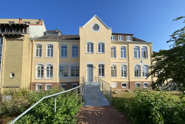 Ein großes altes Haus, dessen Fassade von der Malerei Norbert Bardtke in einem hellen Gelbton gestrichen wurde. Um das Haus herum ist viel Natur zu sehen und eine Art Brücke führt zur Haustür.  