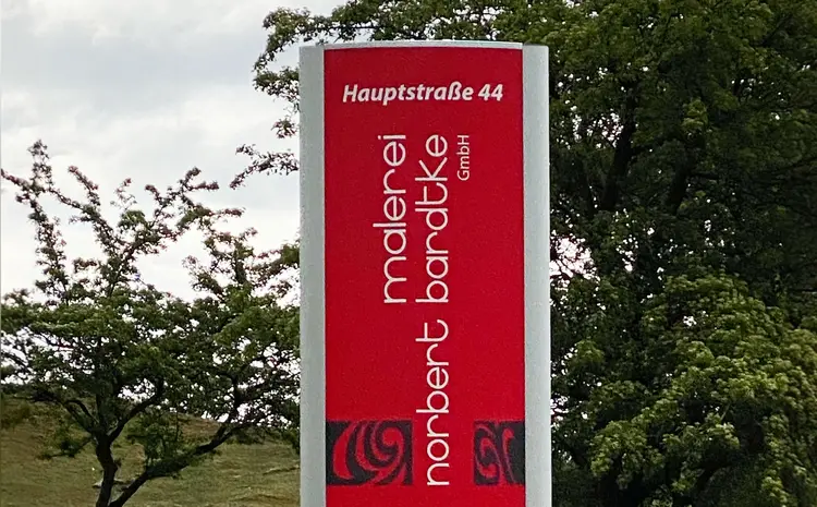 Foto des Markenschildes am Unternehmenssitz der Malerei Norbert Bardtke in Jörl. Ein rotes Schild steht in zwischen einigen Bäumen und trägt den Namen der Malerei sowie die Adresse des Standortes.
